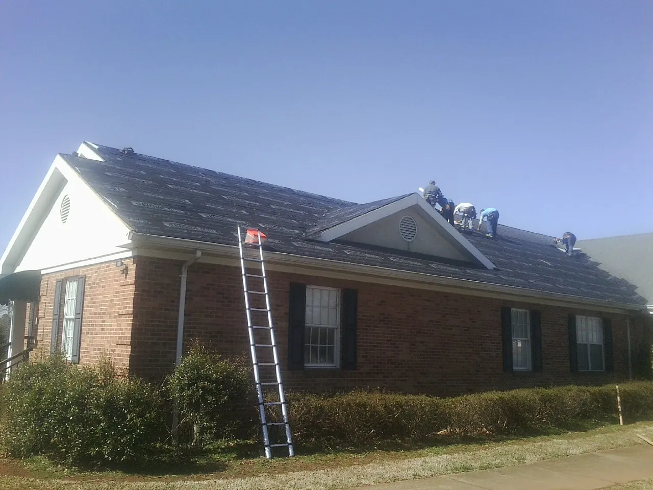 Crew tearing shingles off a brick ranch roof with a ladder set — Metal Roof Repair work by our crew in Choctaw