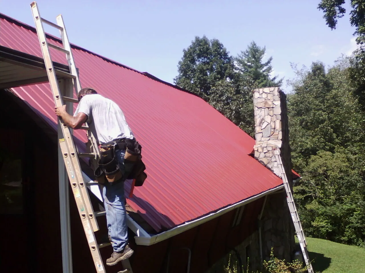Roofer on a ladder beside a red metal roof and stone chimney — Soffit & Fascia Repair by Roofing Brothers in Choctaw